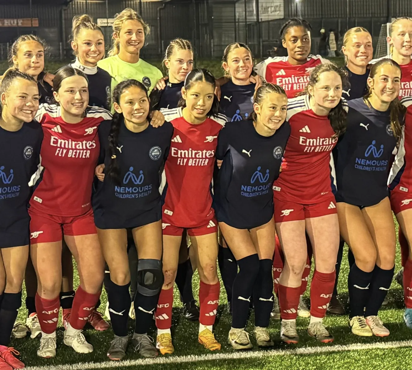Jacksonville FC ECNL girls in navy and Arsenal women’s academy players in red, arm in arm on a floodlit 3G pitch